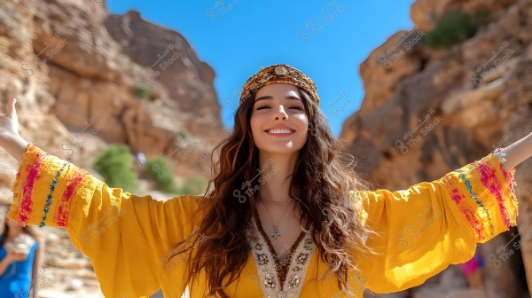 Image of a smiling woman with open arms in a sunny mountainous area. She is wearing an ornate yellow robe and a headband. Behind her are natural rock formations and a blue sky.