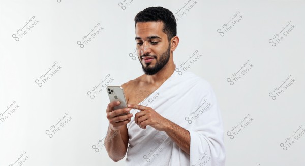 A portrait of a man wearing white ihram clothing and using his smartphone. The person appears engaged with what he sees on the phone\'s screen, with a white background that highlights the features of the image vividly.