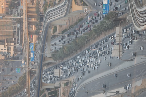 Aerial view of a busy highway in a large city, with numerous cars traveling in both directions during the daytime. The image also features a railway system on an elevated bridge and palm trees lining the sides of the road. Blue signs and signals written in Arabic indicate directions.