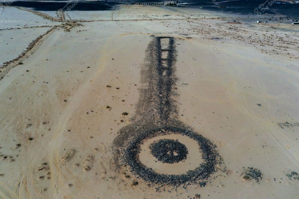 An aerial view of a vast desert area featuring a prominent geometric formation made of black rocks. The formation appears as a long straight line connected to a large circle at its end. The area is surrounded by sand and scattered small rocks. Some sparse desert vegetation and varied terrain are visible on the horizon.