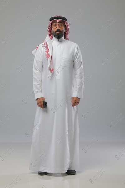 صورة لرجل يرتدي ثوبًا أبيض وغترة حمراء وعقال تقليدي. الرجل يقف في وضعية عمودية ويمسك بيده هاتفًا محمولًا. الخلفية بلون رمادي فاتح. الملابس تشير إلى الزي التقليدي السعودي.