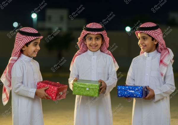The image shows three children wearing traditional Saudi Arabian attire, consisting of a white thobe and a red checkered ghutra with a black agal. They are standing outdoors in the evening, each holding a colored gift box in red, green, and blue. Glowing lights are visible in the background against a night sky.