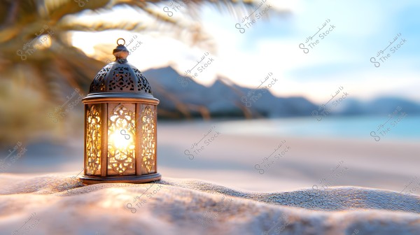 A decorative metal lantern emits light on a sandy beach at sunset. In the background, the sea appears in shades of blue with misty mountains in the distance. Palm leaves are visible, creating a sense of tranquility and peace.