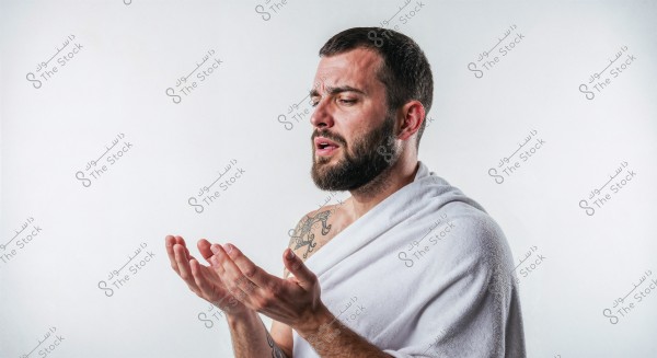 Image of a man wearing a white Ihram, standing with his hands raised in a prayer position. He has a beard and short brown hair, with a tattoo visible on his exposed shoulder. The background is white, highlighting the man\'s features and attire.