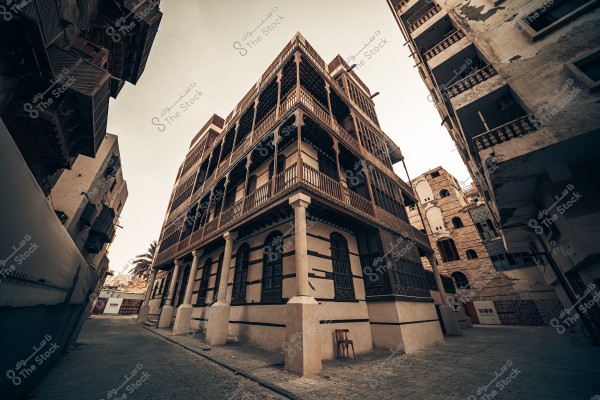 Here’s the translation of your text into English:\r\n\r\n\"A historical image of Jeddah showing a traditional multi-story building at the corner, clearly displaying its unique architectural design with stone and wooden balconies. The surrounding buildings appear slightly worn, giving the area a historical character. The streets are cobblestone, with a wooden chair in front of the central building, and a palm tree visible in the background.\"\r\n\r\nIf you want, I can also make a slightly more **natural, descriptive English version** that reads like a guidebook or historical caption. Do you want me to do that?