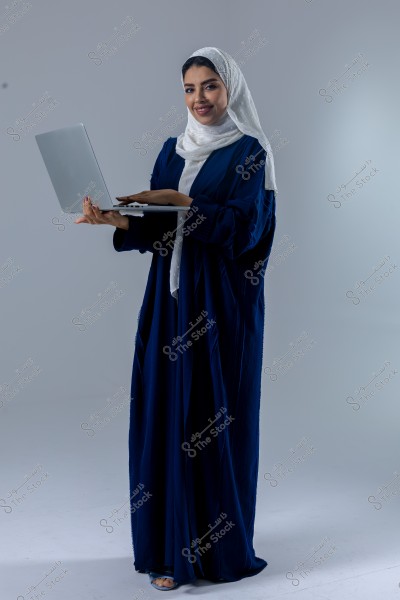A portrait of a woman wearing a blue abaya and a white hijab, holding a laptop. She is standing against a gray background and smiling at the camera. The clothing style is likely of Saudi or Gulf origin.