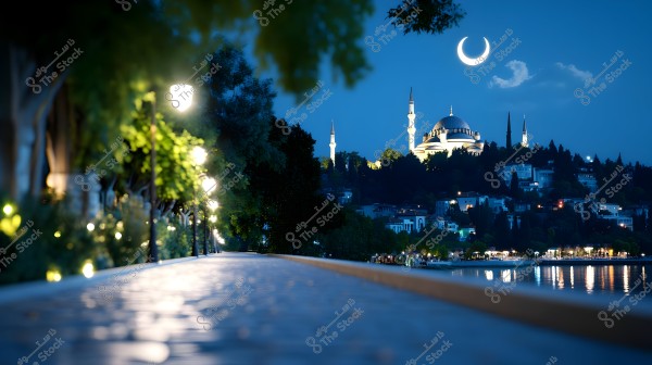 A nighttime view of a large mosque illuminated with white lights, featuring tall minarets, set in the background of a city overlooking a calm lake. The sky is a deep blue with a bright crescent moon above, while streetlights warmly illuminate the pathway by the lakeside, with trees and greenery scattered along the sides.