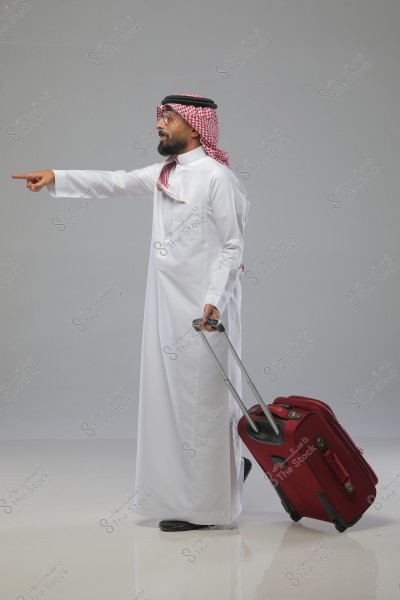 Image of a man wearing traditional white thobe and red ghutra, holding a red suitcase and pointing in a specific direction. The background is grey, suggesting a travel or directional context.