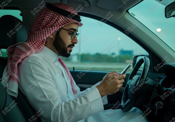 An image of a man sitting inside a car using a mobile phone. The man is wearing a white thobe and a red ghutra with a black agal, indicating traditional Saudi attire. He is sitting in the driver\'s seat, focusing on the phone, with some road scenery visible through the window in the background.