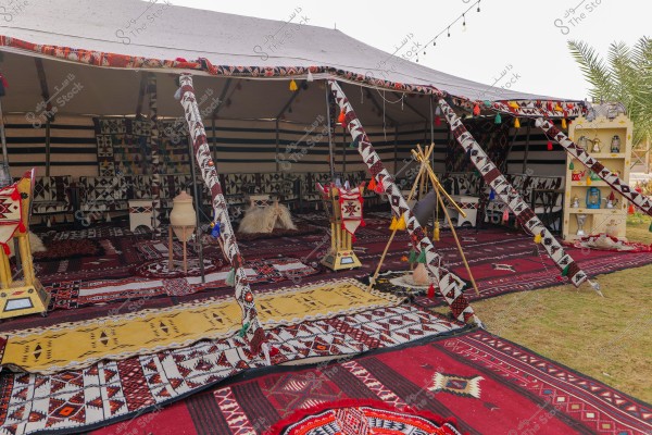 An image of a Bedouin tent decorated with colorful traditional patterns. The tent is made of canvas and open at the front. Red rugs with geometric designs cover the ground. Inside the tent, there are cushions, colorful ornaments, and traditional seating. The interior walls display fabrics with various patterns. Near the tent, there are shelves holding pottery and traditional Arabian decorations.