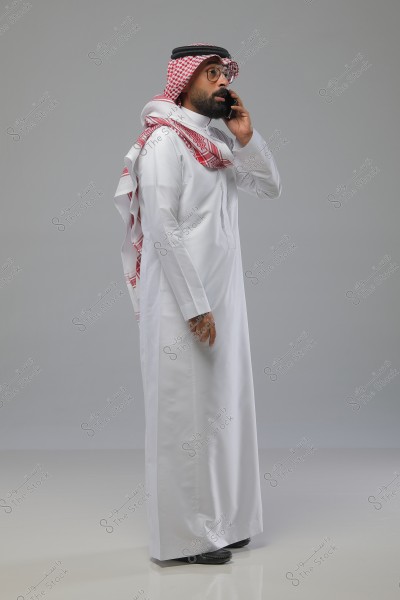 صورة لرجل يقف ويتحدث على هاتف محمول. يرتدي الرجل ثوباً أبيض وغترة حمراء وبيضاء مع عقال أسود, وهو لباس تقليدي شائع في السعودية. لديه لحية ويضع النظارات. الخلفية خفيفة وسادة.