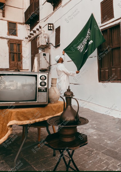 An image of a man wearing a white thobe and kufi, standing next to a traditional building while raising the famous green flag of Saudi Arabia with the Shahada and sword. In the foreground, there is a table with an old TV, a pottery vase, and an Arabic coffee pot, showcasing traditional elements of the setting.