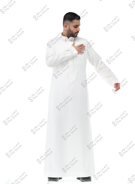 Image of a man wearing a traditional white thobe, standing and looking down at his right sleeve. The thobe indicates a traditional style of clothing common in the Gulf region, possibly from Saudi Arabia.