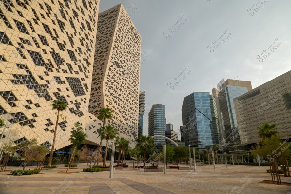 A cityscape featuring a collection of modern buildings with distinctive architectural designs. Some buildings have reflective glass facades, while the large building showcases a lattice design with black and white geometric squares. Trees and palm trees line the foreground plaza, on a calm day with an overcast sky.
