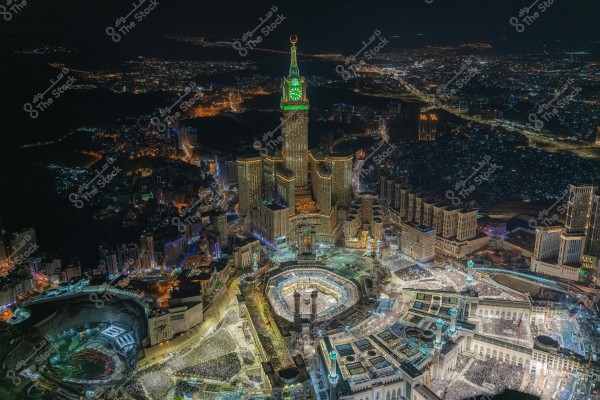 Aerial night view of Mecca, Saudi Arabia, showing the Grand Mosque and the Kaaba surrounded by crowds. The Makkah Clock Tower is illuminated in green, standing prominently in the cityscape, with city lights extending into the surrounding landscape and mountainous background.