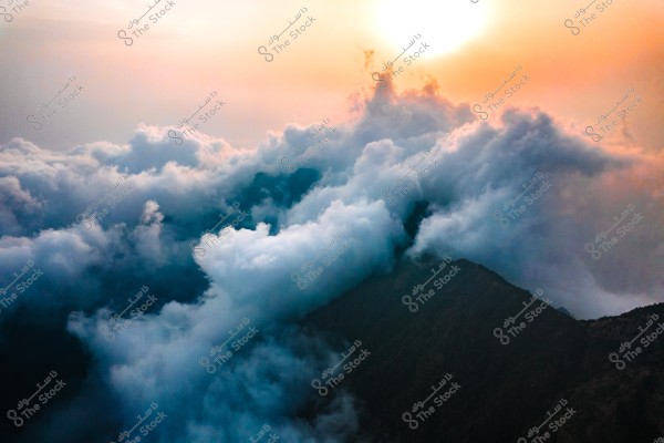 Breathtaking view of thick clouds surrounding mountain peaks at sunset. The orange rays of the sun filter through the illuminated clouds, giving the image a dramatic and tranquil atmosphere. The mountains are barely visible beneath the cloud layer.