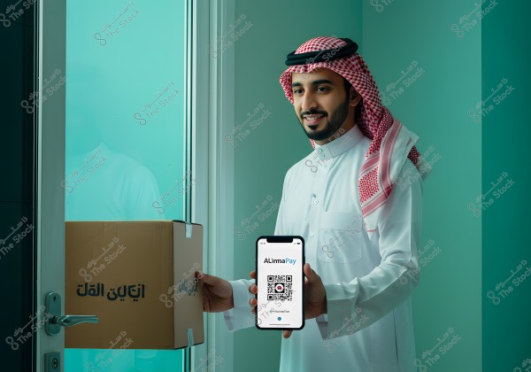 A man wearing a white thobe and a red and white checkered ghutra stands next to a glass door, holding a smartphone displaying a QR code for an app called \"AlimaPay\". In his other hand, he holds a cardboard box with the text \"إيلاك الله\" on it. The background is a light green color.