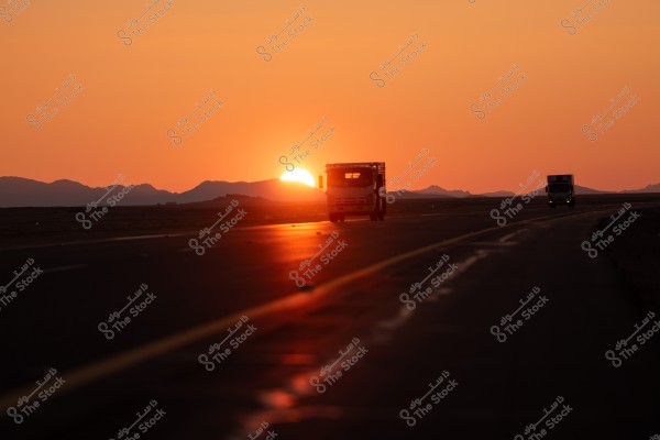 A medium-sized truck is traveling on a highway under a setting sun. The sky is beautifully covered in orange hues, with mountains visible in the distant horizon. Another vehicle is seen on the road on the right side of the image.