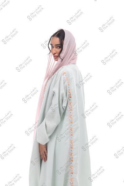 An image of a woman wearing a light blue abaya with floral embroidery on the sides. She is wearing a light pink hijab that partially covers her hair. The woman is looking at the camera with a subtle smile. The background is white and bright.