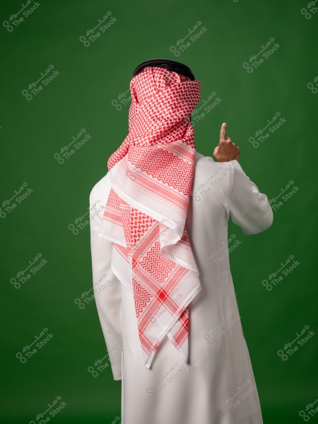 A man wearing traditional Saudi attire in white with a red and white shemagh, pointing upwards, against a green background.