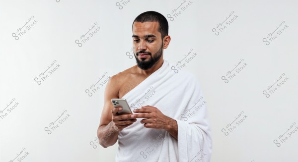 A photo of a man standing against a white background, wearing a white wrap similar to Ihram attire. He holds a smartphone in his hand and looks at it intently.
