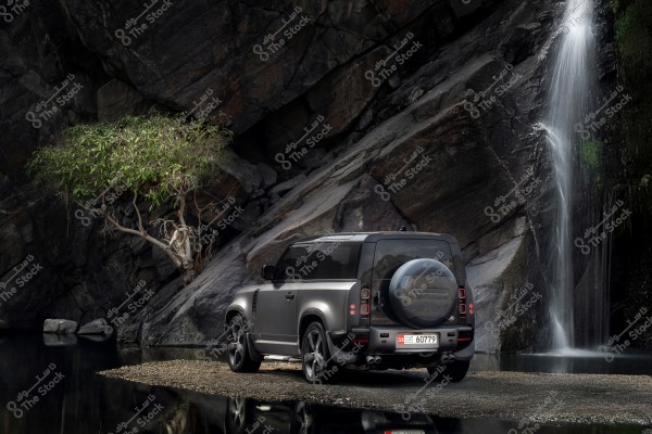 A grey four-wheel drive vehicle is parked by a water\'s edge in front of a small tree and large rocks with a small waterfall in the background. The natural setting creates a serene and picturesque atmosphere.