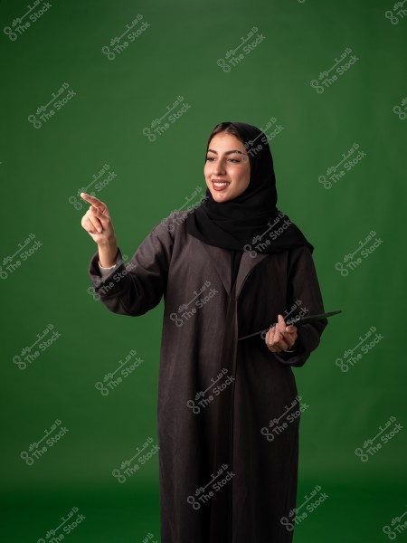 صورة لامرأة ترتدي عباءة سوداء وحجابًا أسود وهي تحمل جهازًا لوحيًا وتشير بإصبعها إلى الجانب. الخلفية خضراء صافية, مما يبرز تفاصيل ملابسها التقليدية. تبدو المرأة مبتسمة ومتعاطفة, مما يضيف الدفء إلى الصورة.