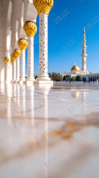 View of part of a mosque with decorative white columns topped with gold, reflecting light on the glossy marble floor. In the background, a golden dome and a minaret are visible, set against a clear blue sky and a row of palm trees.