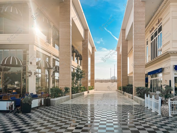 An image of an outdoor plaza between two modern commercial buildings. On the left, there is a café with seating and tables under small awnings. The ground is covered with a black and white checkered tile pattern, and the sun is shining between the buildings with a clear blue sky that has light clouds.