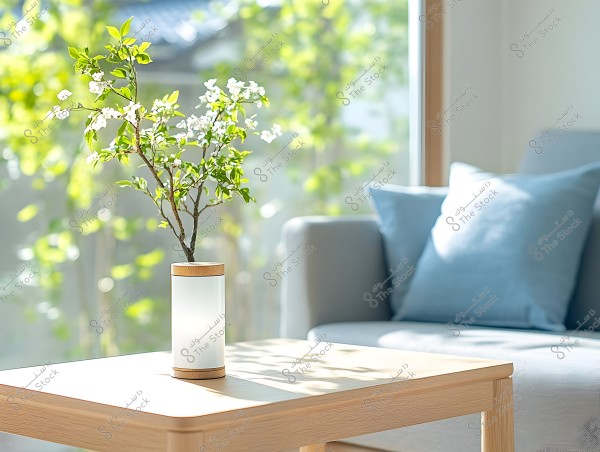 An image of a living room featuring a wooden table with a white vase that has a wooden base, containing small tree branches with white flowers and green leaves. In the background, there is a gray couch with blue cushions, and daylight is streaming in through a large window.