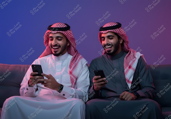 Two men seated on a couch, wearing traditional Saudi attire. The first man is in a white thobe and red checkered shemagh, while the second is in a gray thobe with a red checkered shemagh. Both are looking at their phones with wide smiles, set against a softly lit backdrop in blue and purple hues.