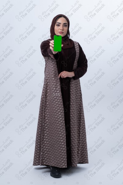 A young Saudi woman wearing an abaya and hijab, holding a green-screen phone