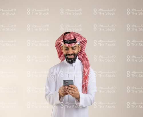 شاب سعودي مبتسم يرتدي الزي السعودي التقليدي يستخدم الجوال، رجل يتصفح هاتفه المحمول