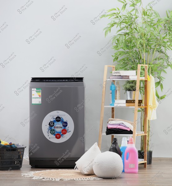 The image shows a silver washing machine in a modern laundry room. Next to it is a wooden shelf with books, a spray bottle, and folded clothes. There is also a green decorative plant. On the floor are a laundry basket with clothes, round cushions, and colorful detergent bottles.