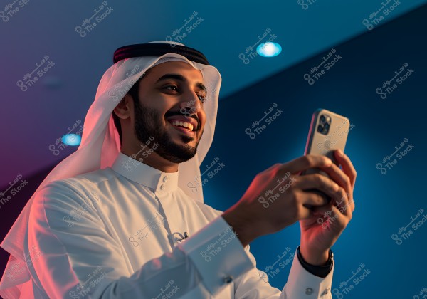 A portrait of a man wearing traditional Saudi attire, including a white thobe, ghutra, and agal, appearing happy while using a smartphone. The ambient lighting adds a modern touch to the scene against a bright blue background.