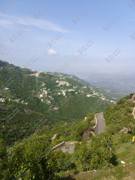 منظر طبيعي لجبل مغطى بالخضرة في يوم مشمس مع طريق متعرج يمر عبر المشهد. تنتشر المباني البيضاء الصغيرة عبر التلال الخضراء, وتظهر السماء الزرقاء الصافية مع بضع غيوم بيضاء في الأفق.