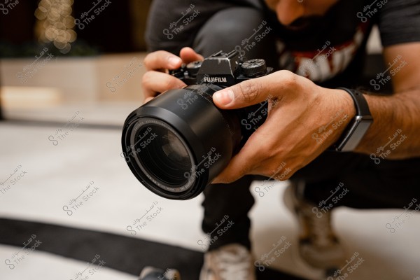 The image shows a person carefully holding a large camera, seemingly preparing to take a photo. The person is dressed casually and is wearing a smartwatch on their wrist. The background features a warm bokeh light effect, suggesting an indoor setting.