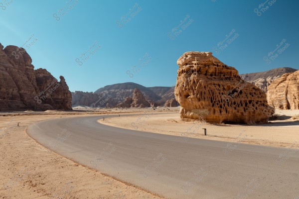An image of a road winding through a desert landscape surrounded by distinctive rock formations. On the right is a large rock with holes and erosion patterns, while rocky formations continue along the left horizon under a clear blue sky. The image highlights the desert environment and the natural sculpting of rocks.