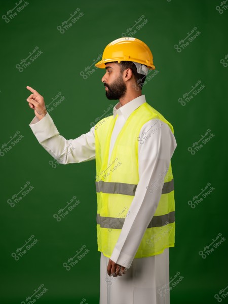 رجل يرتدي سترة سلامة صفراء وخوذة بناء صفراء, يشير بإصبعه في اتجاه معين, مع خلفية خضراء.