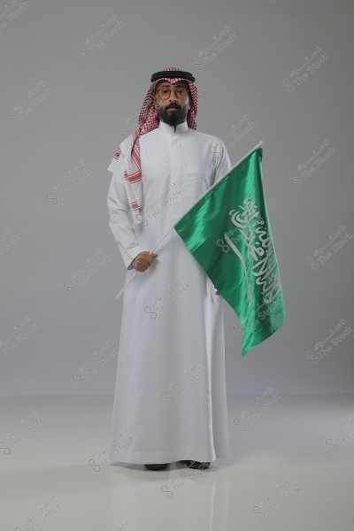 A portrait of a man wearing traditional white attire, including a thobe and a red and white checkered headscarf with a black agal. He is holding the green flag of Saudi Arabia. The man stands against a plain gray background, looking directly at the camera.