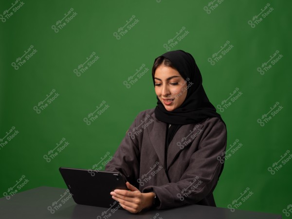 امرأة ترتدي حجاباً وملابس رسمية تجلس أمام شاشة خضراء تستخدم جهازاً لوحياً.