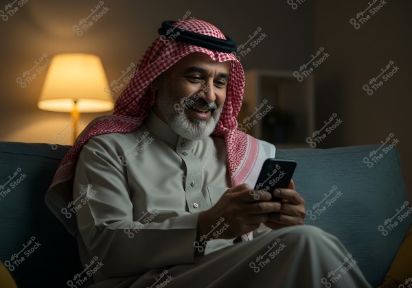 A portrait of a man sitting on a couch, smiling broadly while browsing his phone. The man is wearing traditional Saudi attire, a thobe, and a red and white ghutra. He is seated in a room illuminated by a warm side lamp, creating a cozy and peaceful atmosphere.