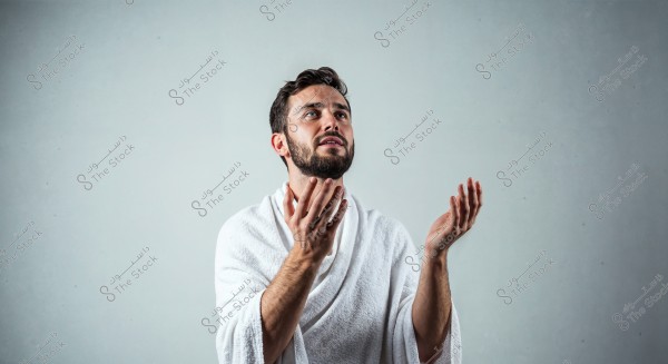 Image of a man wearing a white robe appearing to be in a praying position, standing against a light gray background. The man has a beard and short brown hair, with his hands raised as if in supplication or worship.