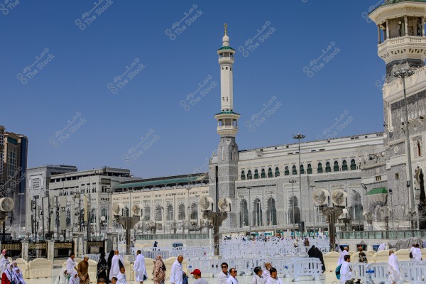 The image shows the courtyard of the Grand Mosque in Mecca, Saudi Arabia, with several pilgrims wearing white ihram garments. The famous minaret and the mosque with its ornate stone facade are in the background, set against a clear blue sky.