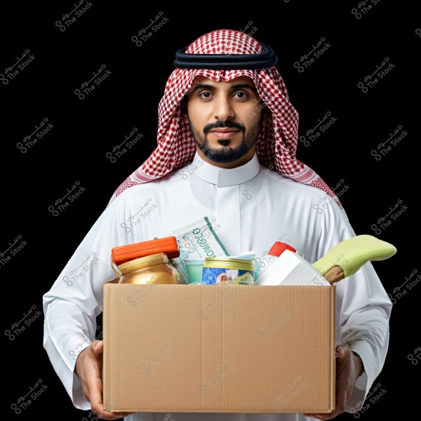 A portrait of a man wearing a traditional Saudi thobe and keffiyeh, standing and holding a cardboard box filled with groceries, such as a glass jar, metal cans, and other containers. The background is black.