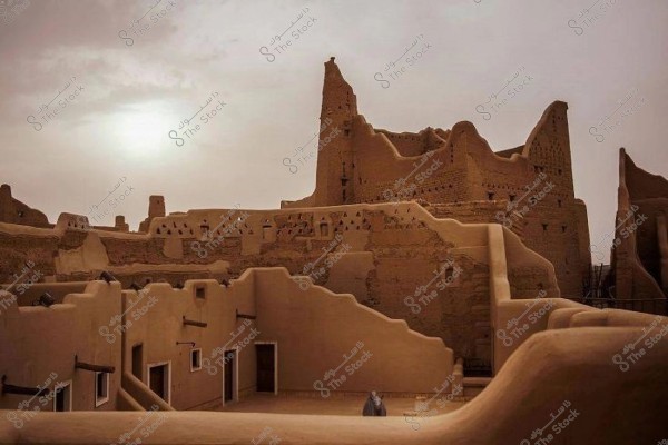 A scene of a historic mud-brick fortress in the evening with the sun setting in the background. The fortress consists of high walls built from mud bricks with geometric shapes and ancient markings. The sky above the fortress is cloudy, adding a historical and mysterious feeling to the place.