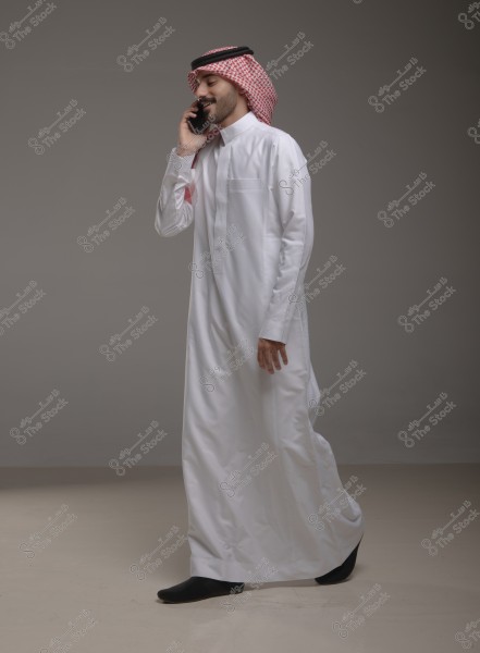 Image of a man wearing traditional Saudi attire, consisting of a white thobe, a red and white checkered shemagh, and a black agal. The man is speaking on the phone and appears to be smiling. The background is gray.