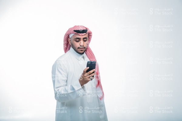 رجل سعودي يرتدي الزي الرسمي السعودي على خلفية بيضاء ، يمسك الجوال ، يستخدم أحد تطبيقات الآيفون ، يتصفح الإنترنت ، يستخدم وسائل التواصل الاجتماعي