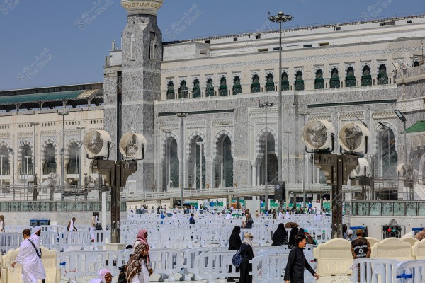 An image of a bustling area near the Grand Mosque in Mecca, Saudi Arabia. The picture shows many people wearing white Ihram garments, while others wear black abayas. The buildings in the background feature marble cladding and large arched windows. Large fans are distributed in the courtyard to help with ventilation.