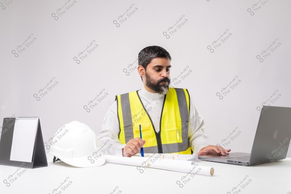 A photo of a man sitting at a desk working on a laptop. He is wearing a yellow reflective vest over a traditional white thobe. Next to him are a white helmet, a notebook, and architectural blueprints. He appears to be working in the engineering or construction field.